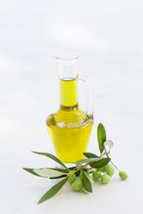 Olive oil in glass bottle with fresh olives on branch on white table.