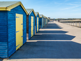 Blue and yellow wooden beach huts casting long shadows in the sunshine along the promenade at the coast of Minnis Bay, Kent, UK