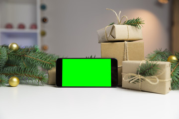 Smart phone with green screen surrounded by Christmas decorations. Holiday and christmas concept. Wrapped christmas gifts.