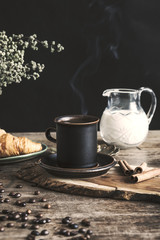 The vintage and stylish composition in kitchen interior with a cup of coffee, milk and croissant. Winter and autumn concept of morning breakfast. Black background.