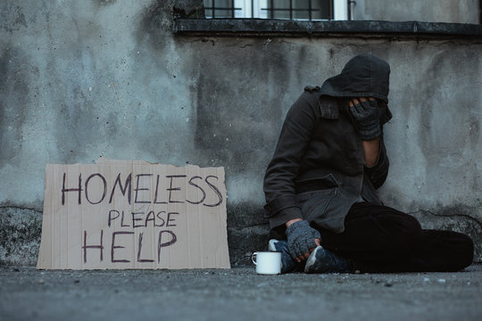 Homeless man sitting on the street in the shadow of the building and begging for help and money. Problems of big modern cities.