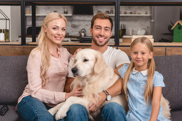 parents with daughter and dog sitting on couch at home and looking at camera