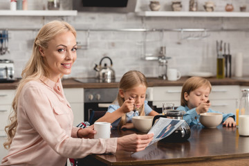 Obraz premium smiling mother having cup of coffee while sitting at kitchen with kids during breakfast and looking at camera