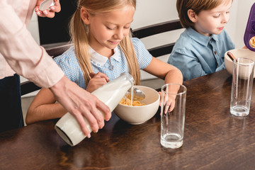 cropped shot of mother pouring milk into cereal for kids during breakfast