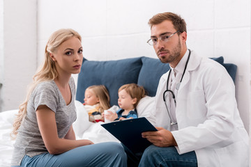 Fototapeta premium pediatrician and mother looking at camera while sick kids lying in bed on background
