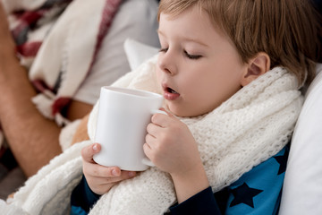 close-up portrait of little kid in scarf with cup of hot drink sitting in bed