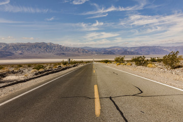View of a straight section of California State Route 190 in Death Valley National Park with arid desert on the road-side and the Sierra Nevada mountains at the horizon against a blue sky with clouds
