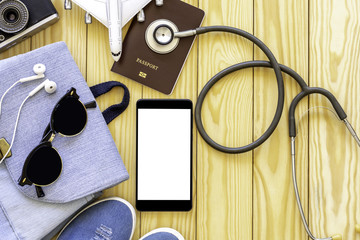 Mock up image of mobile smart phone with blank white screen, passport, camera, airplane model,  sunglasses and medical stethoscope isolated on wooden background. Technology and travel concept