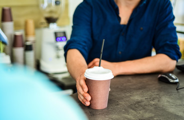 Fototapeta na wymiar Hand gives cup to client visitor. Man receive drink at bar counter. Cappuccino or cacao with straw. Served in paper cup coffee to go. Hand of barista at bar serve coffee for client. Enjoy your drink