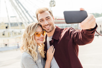 smiling couple in autumn outfit taking selfie with smartphone in city