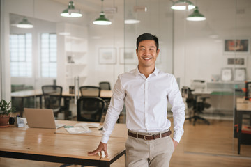 Young Asian businessman standing in an office smiling confidently - obrazy, fototapety, plakaty