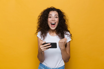 Happy cute young woman posing isolated over yellow background play games by mobile phone.