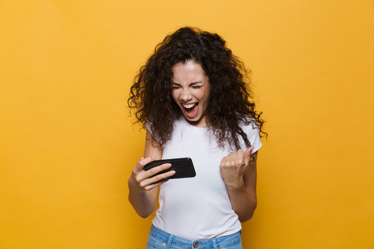 Happy cute young woman posing isolated over yellow background play games by mobile phone.