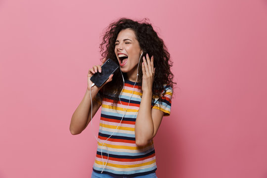 Image of cheerful woman 20s with curly hair singing while holding smartphone like microphone and listening to music via earphones, isolated over pink background