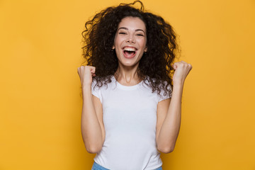 Image of young woman 20s with curly hair yelling and clenching fists, isolated over yellow...