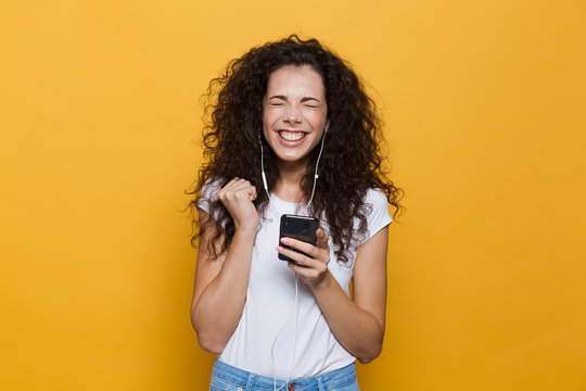 Image of happy woman 20s with curly hair holding smartphone and listening to music via headphones, isolated over yellow background