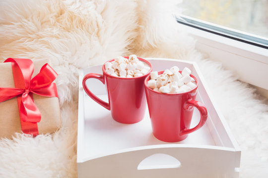 Two red cup of hot chocolate with marshmallow on white windowsill with furskin for rest. Holiday. Romantic.
