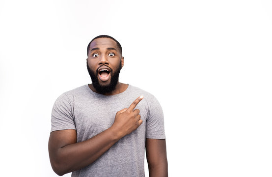 African American man grins at camera, indicates copy space for tex or logo, advertises something. Surprised man points with fore fiinger, shocked expression, isolated over white background. Look here!