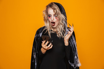 Pretty woman wearing black costume and halloween makeup holding smartphone, isolated over yellow...