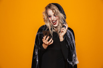 Scary wizard woman wearing black costume and halloween makeup holding smartphone, isolated over...