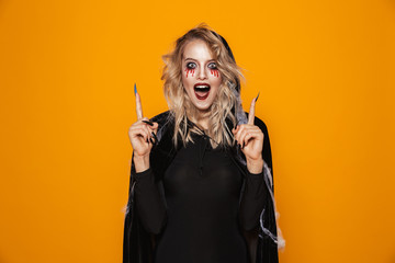 Amazing woman 20s wearing black costume and halloween makeup pointing fingers upward, isolated over...