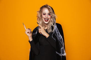 Young woman 20s wearing black costume and halloween makeup pointing fingers aside, isolated over...