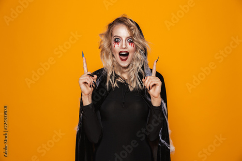 Amazing woman 20s wearing black costume and halloween makeup pointing fingers upward, isolated over yellow background