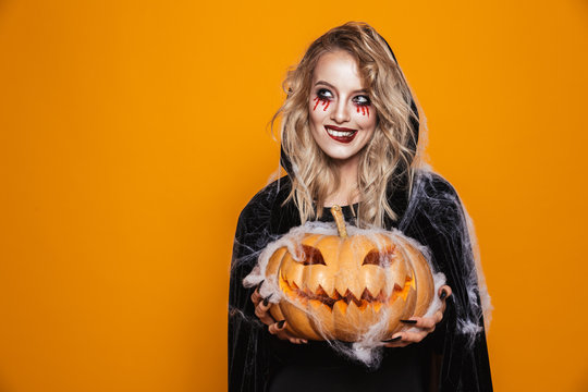 European witch woman wearing black costume and halloween makeup holding carved pumpkin, isolated over yellow background