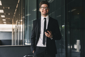 Portrait of attractive businessman dressed in formal suit standing outside glass building, and holding mobile phone