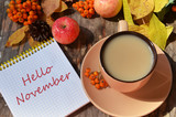 Tag with words hello november on the notebook cup of coffee lying on the books with dry yellow maple leaves and everlasting flowers on background