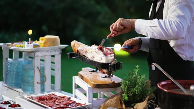 Caterer on the event party. Food service worker who cuts a slice of ham, of excellent quality, with the knife according to tradition outdoors in the restaurant. Concept of ham, food, Italy and Spain