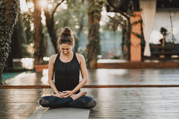 Fototapeta premium Concentrated girl sitting in lotus pose and meditating or praying. Young woman practicing yoga alone on wooden deck in tropical island