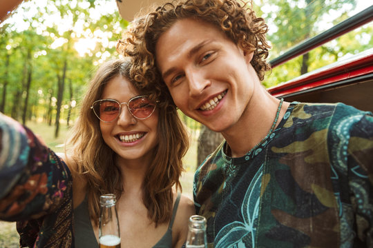 Photo of beautiful hippie couple man and woman smiling, and taking selfie in forest near retro minivan