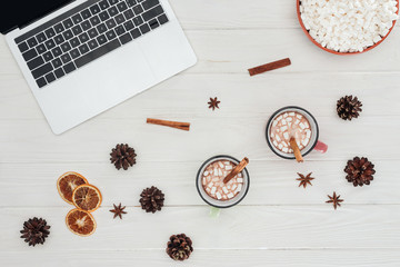flat lay with laptop, mugs of hot chocolate and cinnamon sticks with marshmallows on wooden table