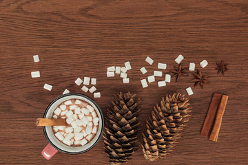 top view of cup with hot chocolate, marshmallows, pine cones and cinnamon sticks on wooden table