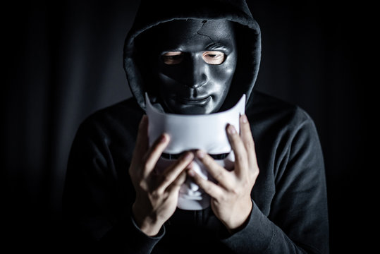Mystery hoody man wearing broken black mask holding white mask. Anonymous social masking. Major depressive disorder or bipolar disorder. Halloween concept