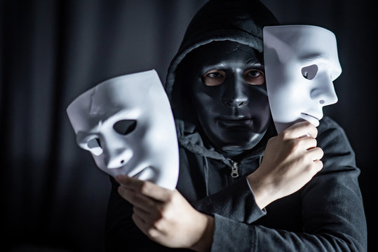 Mystery hoody man wearing black mask holding two white masks in his hand. Anonymous social masking. Major depressive disorder or bipolar disorder. Halloween concept