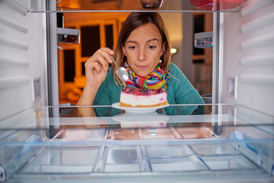 Woman eating gateau while standing in front of fridge. Picture taken from the inside of fridge.