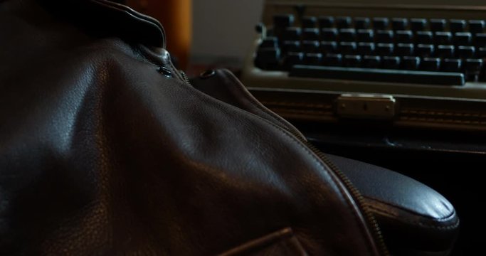 A shot of an old leather jacket hanging off of a desk chair, that racks focus to an antique suitcase typewriter. It has a shallow depth of field and moody lighting.