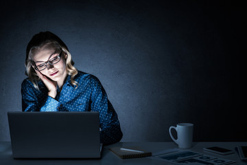 Attractive blonde working on laptop in dark office. Mixed media