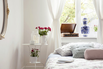 Red roses on table by a bed side in a pastel bedroom interior with a sunny window and a vintage gramophone. Real photo.