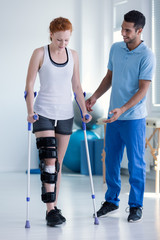 Physiotherapist helping woman with stiffener on the leg walking with crutches