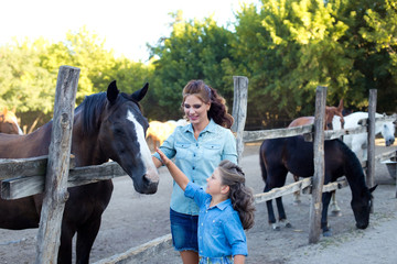 A small smiling girl with mother with curly hair dressed in jeans staying and feeding horses at the stable