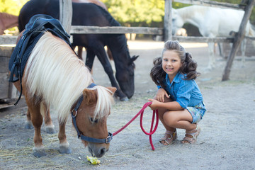 A small smiling girl with curly hair dressed in jeans feeding a pony at the stable