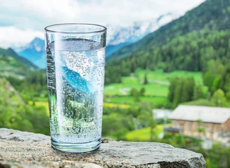 Foto op Plexiglas Glas water op de steen. Wazig sneeuw bergen toppen en groene bossen op de achtergrond. © volff