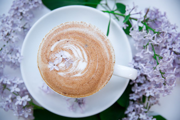 A cup of cappuccino top view is on the branches of lilac, on the foam lie the flowers. Чашка капучино вид сверху стоит на ветках сирени, на пенке лежат цветки.
