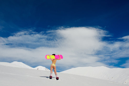 Rear view of young sexy girl dressed in a bright yellow swimsuit with a snowboard on her shoulders