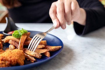Closeup image of a woman's hand using fork to eat fried chicken and french fries in restaurant