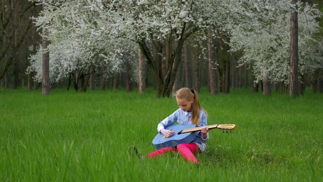 Young beautiful little blonde girl sitting in a wonderful place plays guitar. on background of a blossoming tree green grass in the spring forest