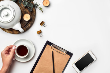 Young woman right hand writing on blank notebook on white table with tea cup, smartphone, and kettle beside in morning time